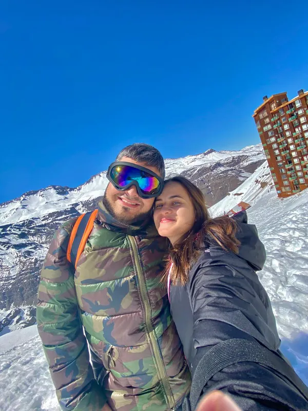 Selfie do casal na neve, com montanhas e céu azul ao fundo.