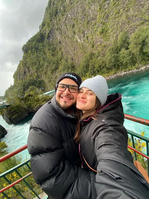 Isabel e Heins de gorro e casaco em um mirante, com um rio azul-turquesa e uma montanha ao fundo.