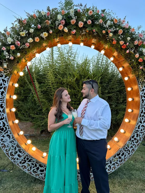 Isabel e Heins, bem-vestidos, brindando diante de um arco de flores iluminado.