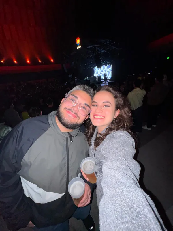 Selfie de Isabel e Heins com copos de cerveja no show do Pedro Sampaio, com o palco ao fundo.