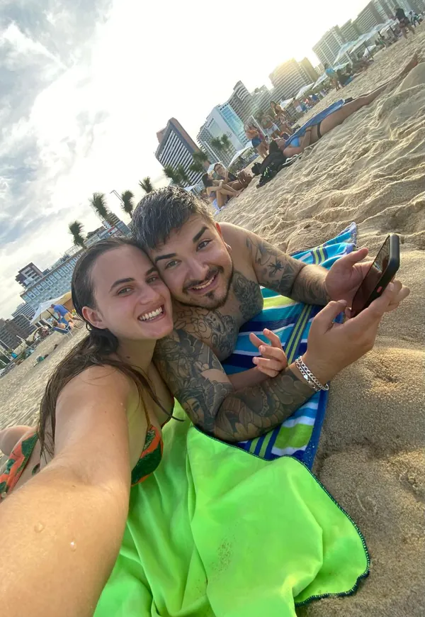 Isabel e Heins deitados na areia, com prédios e coqueiros ao fundo.